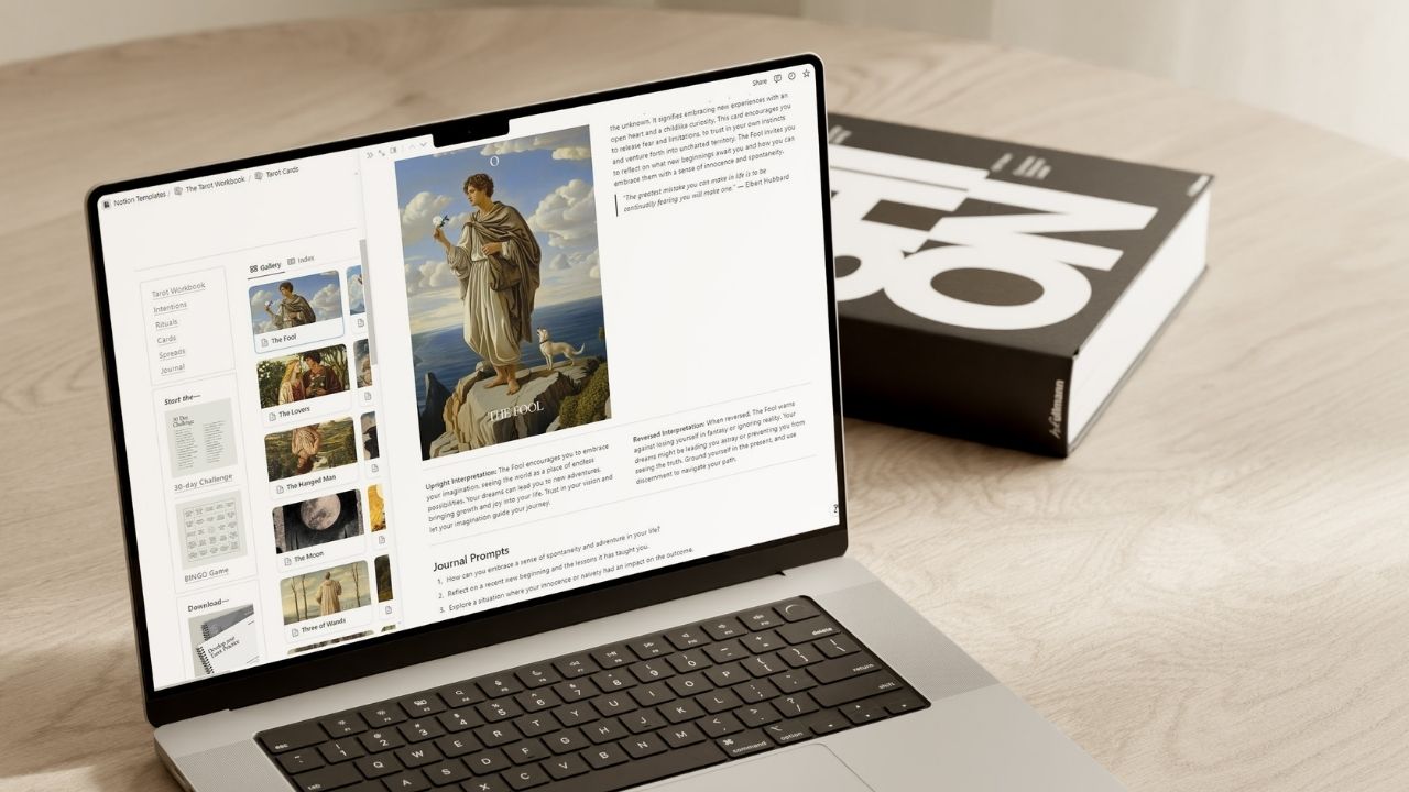 Open laptop displaying a Tarot Workbook in Notion, with a card library and detailed description of 'The Fool' tarot card, including journal prompts and upright/reversed interpretations. A book is placed beside the laptop on a wooden table.