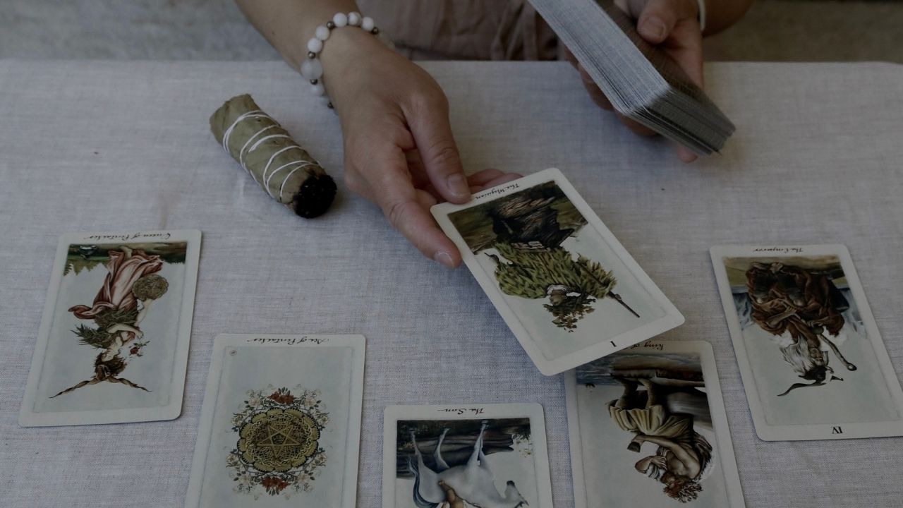 A hand holding tarot cards during a reflective tarot reading session, featuring The Magician, Queen of Pentacles, and other cards. A bundle of bayabas rests beside the cards on a cloth-covered table, symbolizing mindfulness and self-reflection.