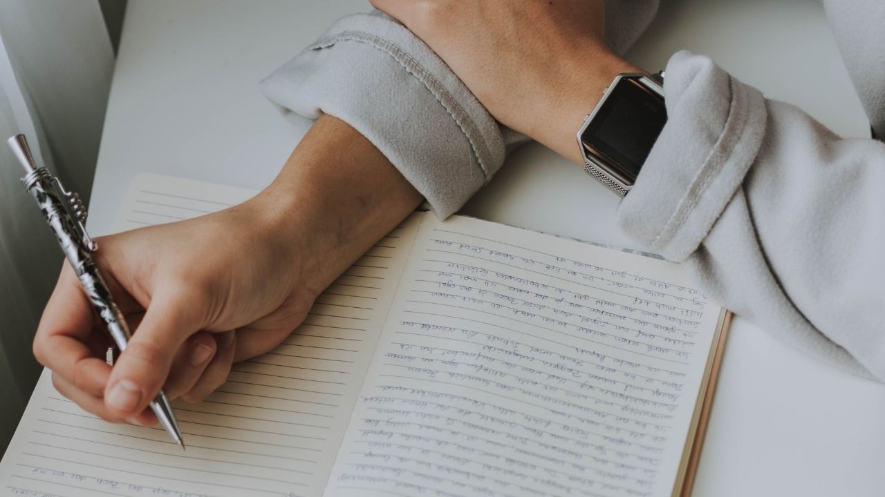 Close-up of a person writing in a lined journal with a silver pen, wearing a smartwatch, focusing on self-reflection through writing.