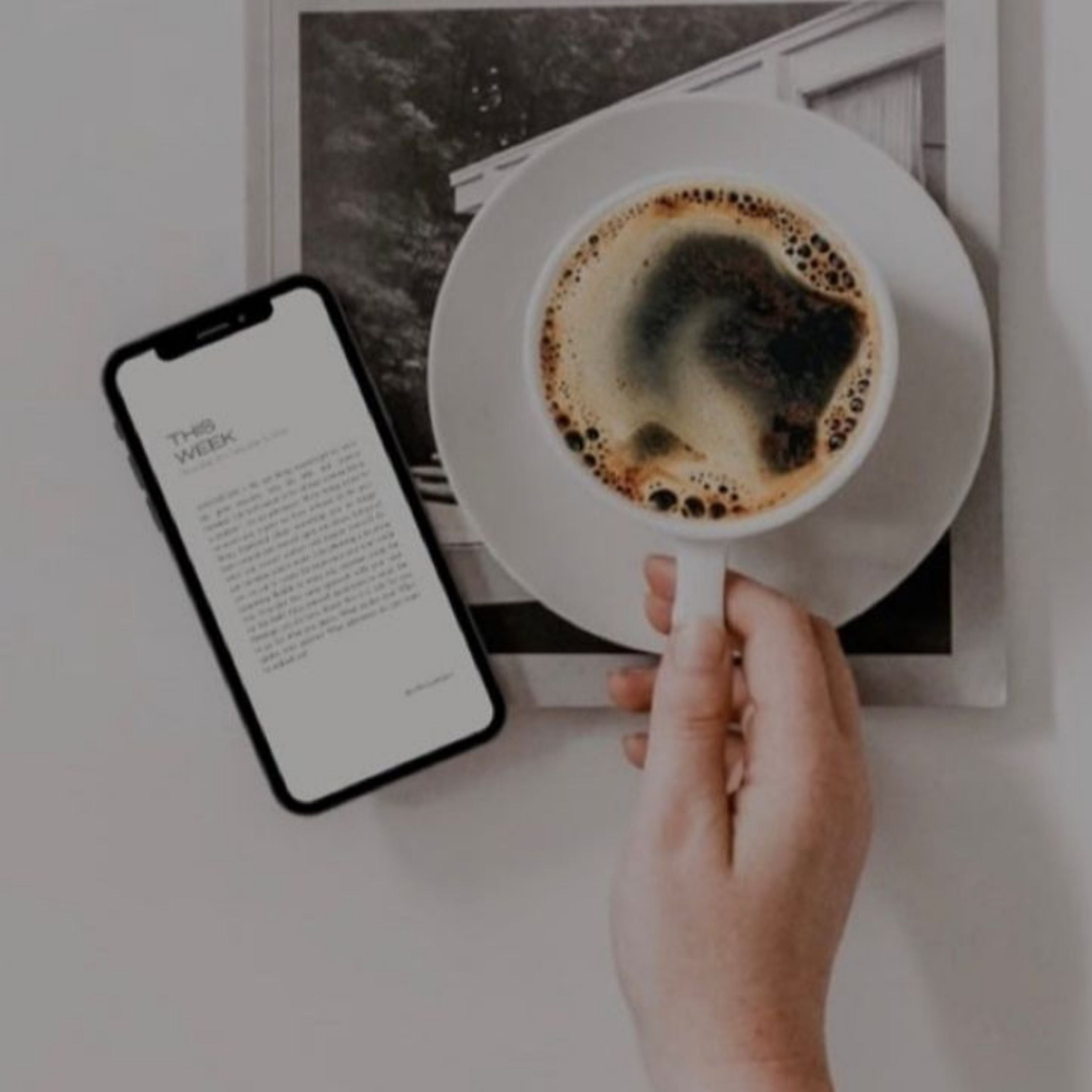 Flat lay of a hand holding a cup of coffee next to a smartphone displaying a weekly newsletter on a minimal table setting.