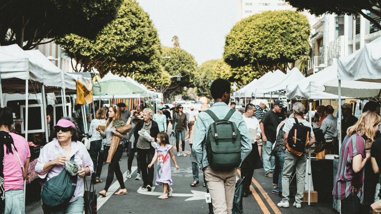 A bustling outdoor market filled with shoppers, vendors, and families walking among vendor stalls. People are interacting and exploring goods in a tree-lined street. Photo by Dane Deaner on Unsplash.