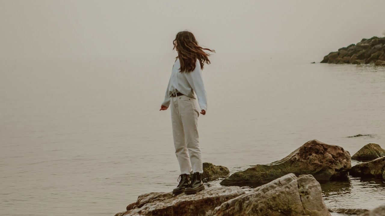 A woman standing on a rocky shore, gazing out over a calm sea, symbolizing introspection and personal growth. This image represents the inward journey with tarot and self-reflection. Photo by Jasmin Chew on Unsplash.