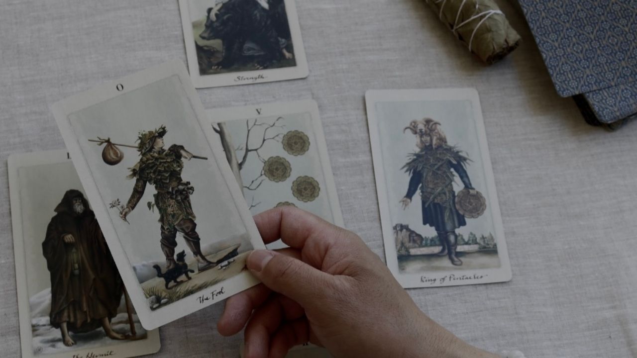 A hand holding "The Fool" tarot card in a tarot card reading setup, surrounded by other cards including "King of Pentacles," "Strength," and "The Hermit." A sage bundle and tarot deck are placed in the background, creating a serene, reflective atmosphere. Featured cards are from the Pagan Otherworlds by Uusi.